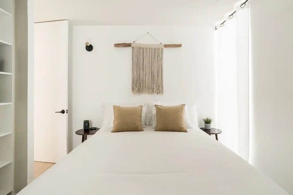 A minimalist bedroom with a large bed, two bedside tables, a hanging macrame wall decor, and a small potted plant.