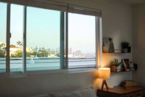 A room with a large window, a lamp on a bedside table, and a shelf with books and decorative items.