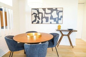 A dining area with a round table, four chairs, a wall-mounted abstract painting, and a small side table with a vase.