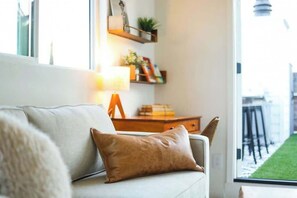 Ein Wohnzimmer mit einer Couch, einer Lampe, einem Holztisch und Blick auf eine grüne Außenanlage durch ein Fenster.