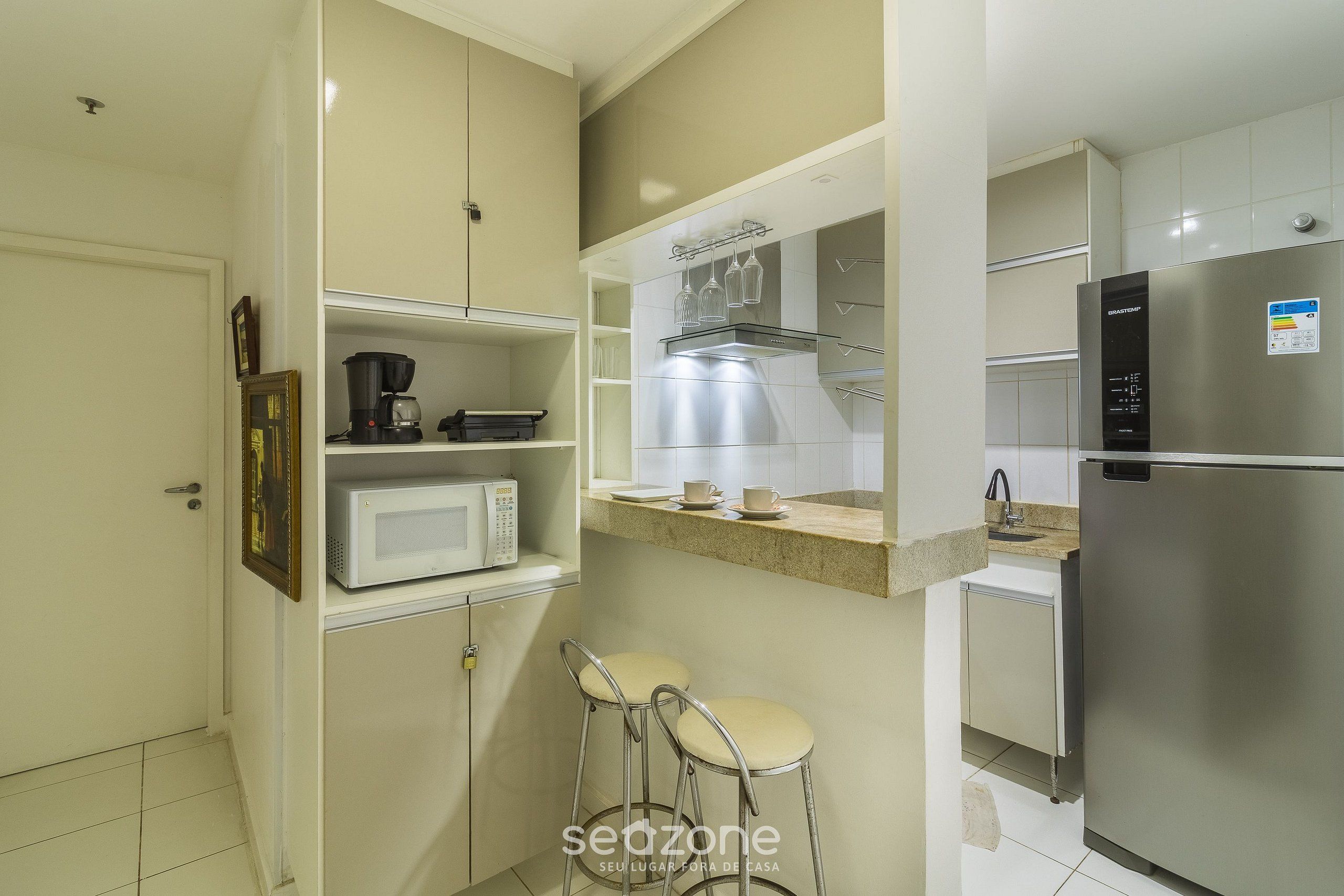 A compact kitchen with a microwave, coffee maker, and stainless steel appliances. There are two bar stools and a small countertop with a sink.