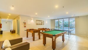 Een ruime kamer met een pooltafel, een tafelvoetbaltafel en een biljarttafel. Er hangt een brandblusser aan de muur en een raam met uitzicht op palmbomen.