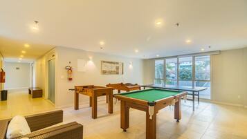 A spacious room with a pool table, a foosball table, and a billiards table. There is a fire extinguisher on the wall and a window with a view of palm trees.