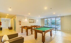 Ein geräumiger Raum mit Billardtisch, Kicker und Pooltisch. An der Wand hängt ein Feuerlöscher und durch das Fenster bietet sich der Blick auf Palmen.