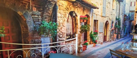 A narrow street with stone buildings, a bed with blue and yellow bedding, and a small table with a remote control.