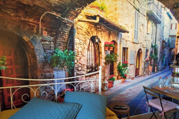 A narrow street with stone buildings, a bed with blue and yellow bedding, and a small table with a remote control.