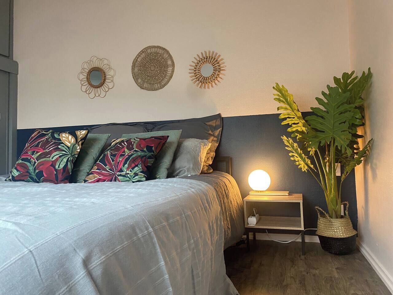 Een slaapkamer met een bed, kussens, een nachtkastje met een lamp en een potplant.