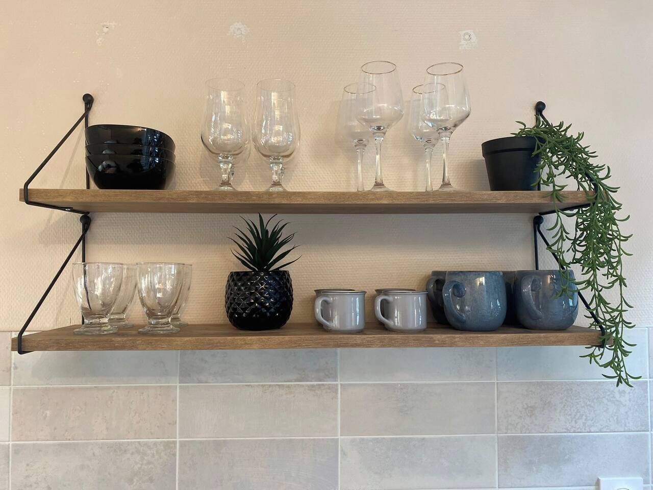 Dos estantes de madera con cristalería, una piña y tazas de cerámica frente a una pared de azulejos.