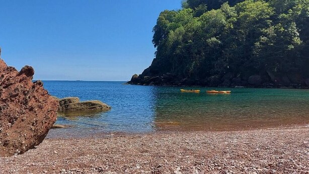 Una playa rocosa con aguas claras, una gran formación rocosa y un acantilado con vegetación densa.