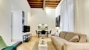 A modern living room with a beige sofa, a coffee table, and a TV mounted on the wall. There is a dining area with a table and chairs, and a staircase leading to an upper level. - TREVI CHIC HOUSE CENTER OF ROME (Roma)