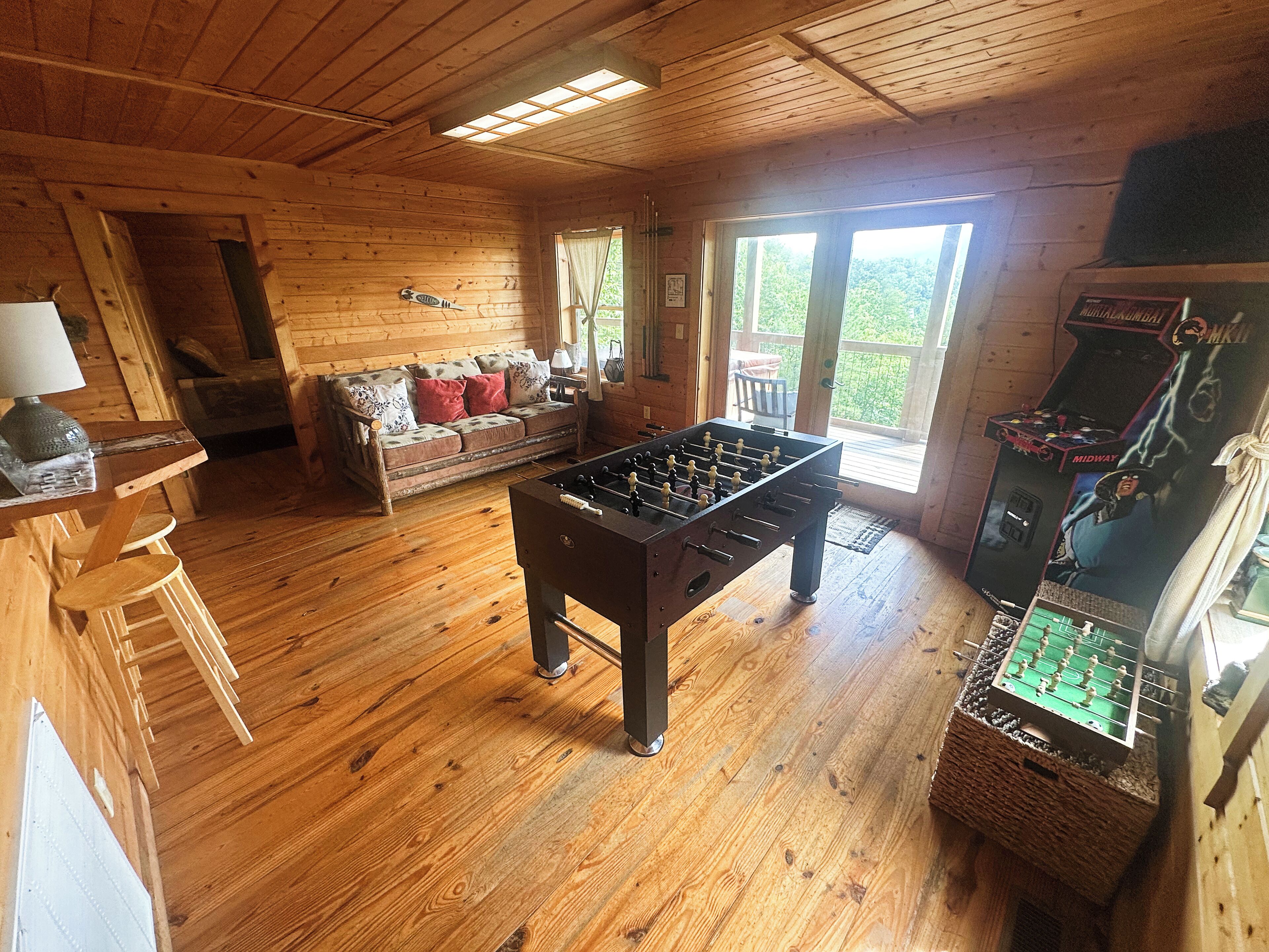 Ein gemütliches Zimmer mit Holzdielen, einem Kicker, einer Couch mit Kissen und einem Fenster mit Vorhängen.