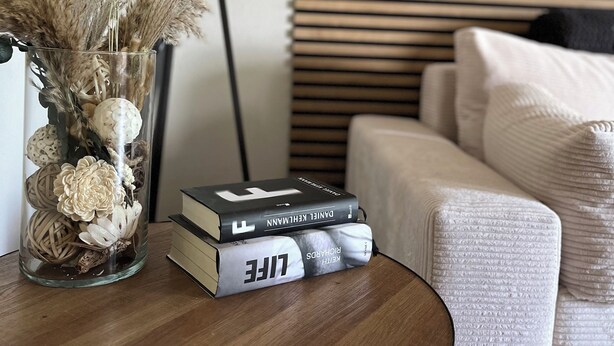 Une table ronde en bois sur laquelle se trouve un vase en verre contenant des plantes séchées et une pile de livres. Une lampe de sol noire est placée à côté de la table, et un canapé avec des coussins blancs se trouve à l’arrière-plan.