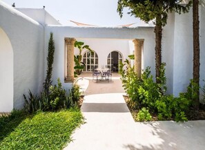 A courtyard with a concrete pathway leading to a seating area with chairs and a table, surrounded by greenery and columns. - Dar Jbel Hammamet (Hammamet)