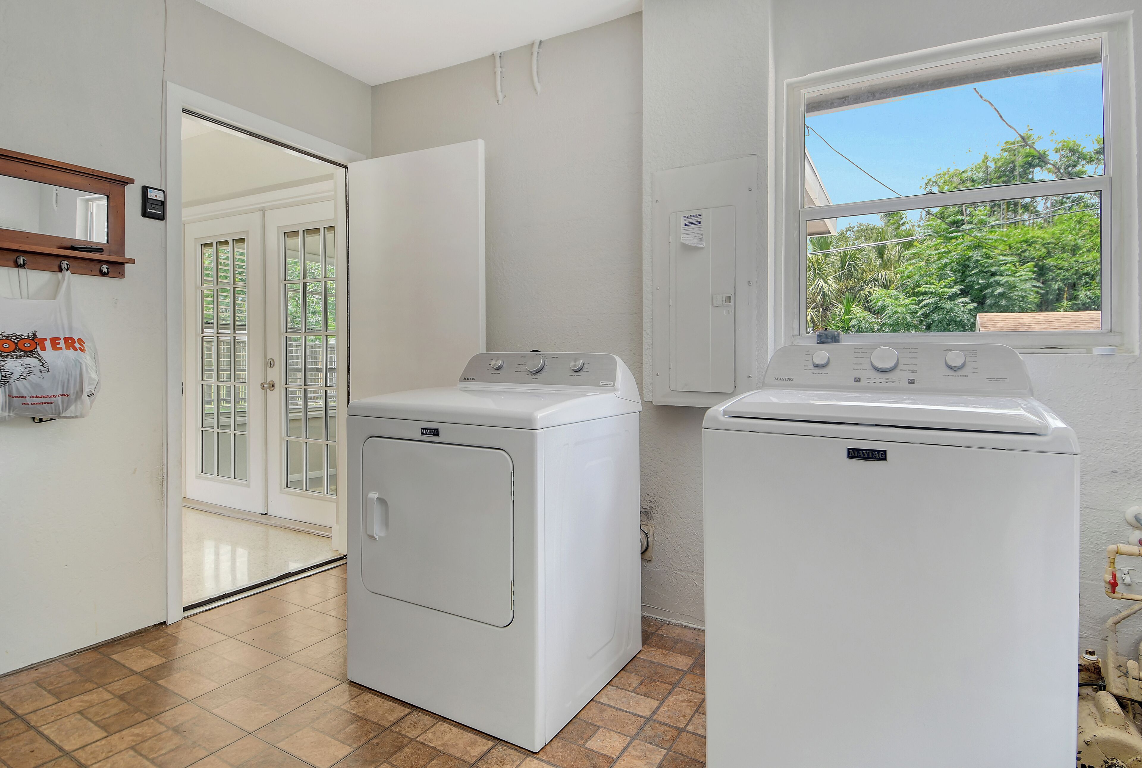 A laundry room with two white Maytag washers and dryers, a wooden shelf with a mirror, and a window with a view of greenery.