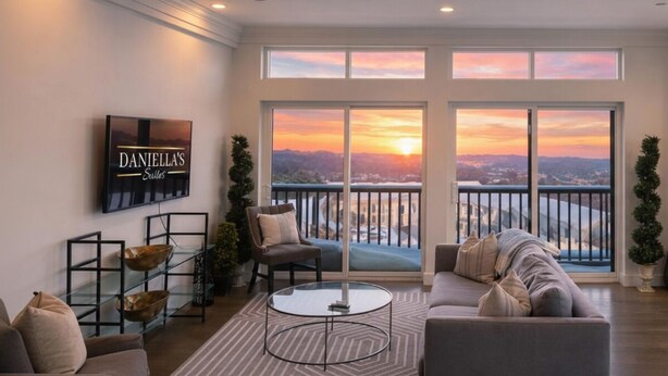 A spacious living room with a large window overlooking a scenic view, a sofa, a coffee table, and a TV mounted on the wall. - Daniella's Suites (Peabody)