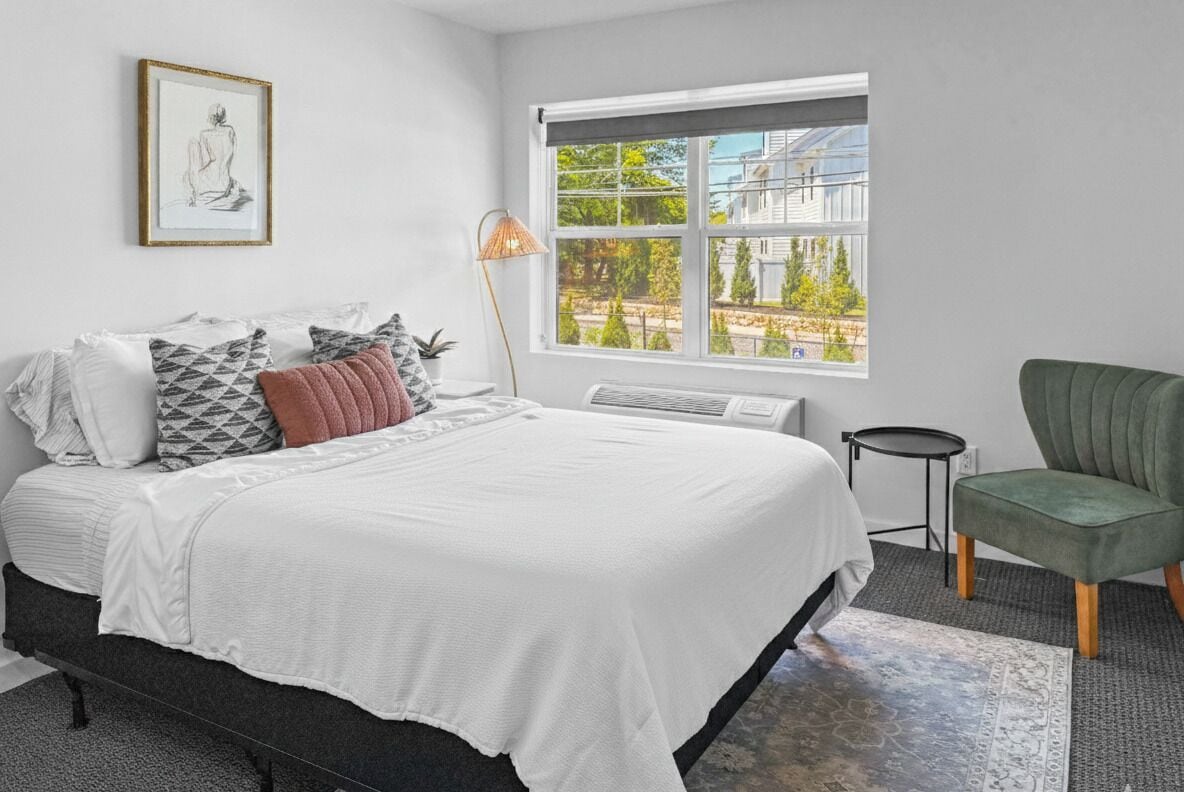 A bedroom with a bed, a side table, a chair, a window with a view, and a framed picture on the wall.
