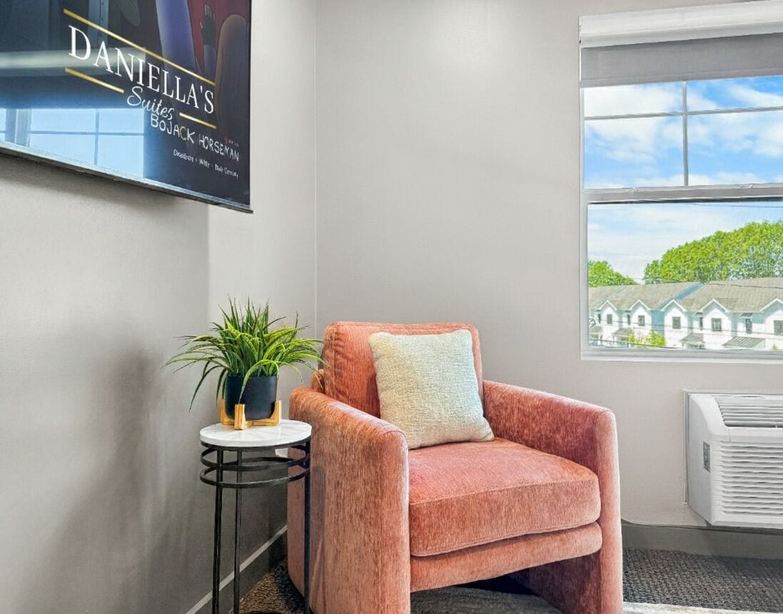 A room with a plush pink armchair, a small round table with a plant, and a large window with a view of houses.