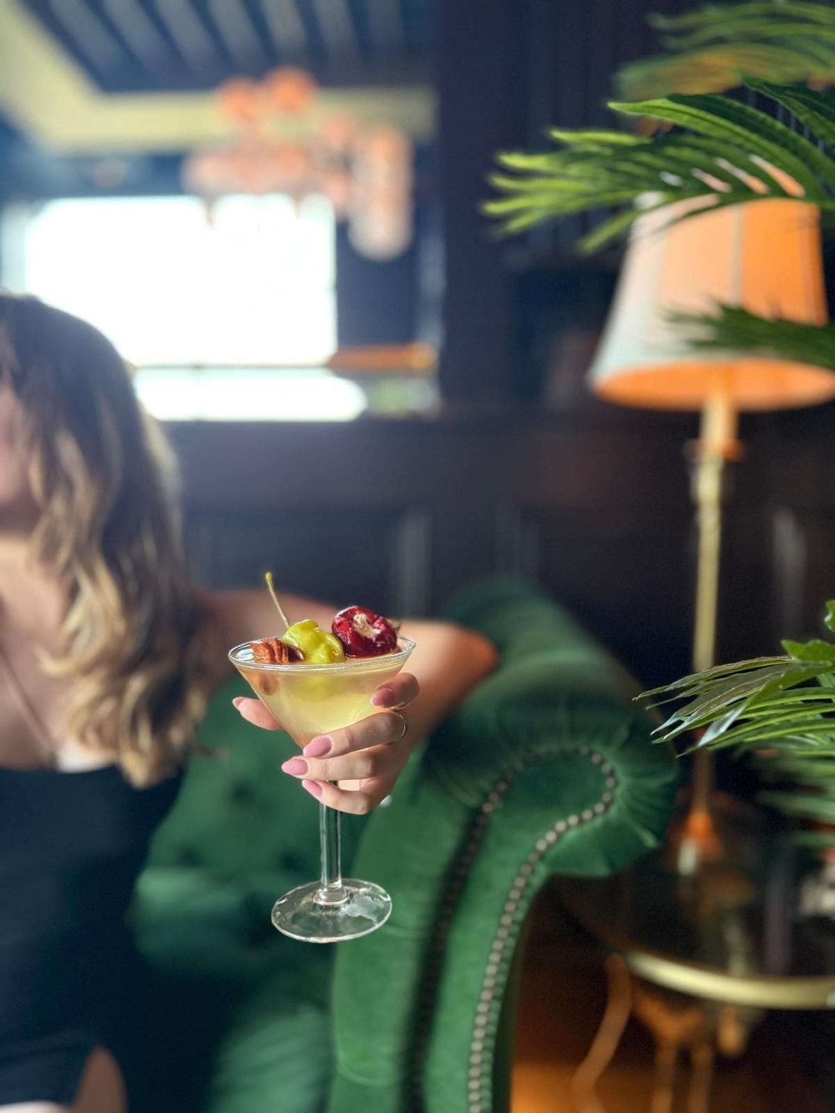 Iemand houdt een cocktail met fruitdecoratie vast, zittend op een groene bekleden stoel in een kamer met een lamp en een plant.