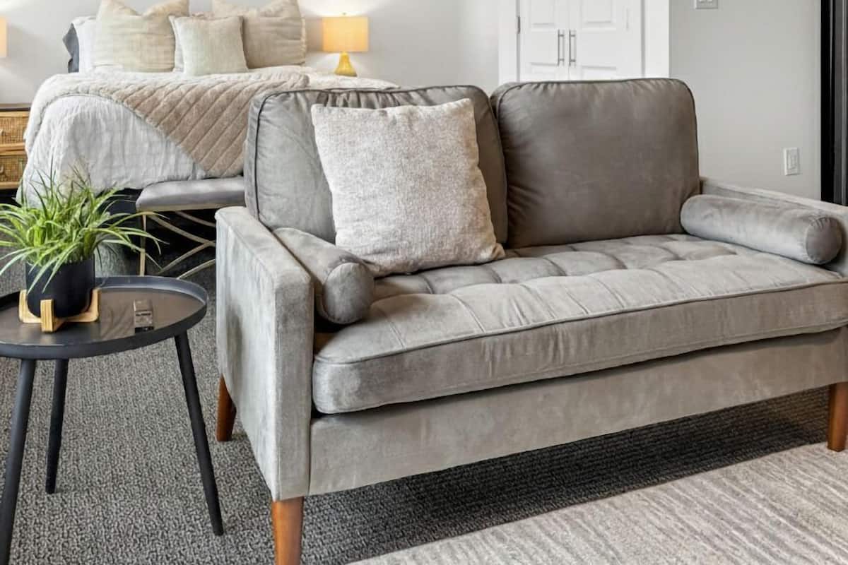 Ein modernes Wohnzimmer mit einem grauen Sofa, einem kleinen runden Tisch mit einer Pflanze und einem Bett mit weißem Kopfteil.