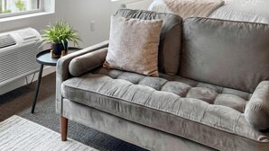 A modern bedroom with a grey sofa, a bed with multiple pillows, a bedside table with a lamp, and two windows with blinds. - Daniella's Suites (Peabody)