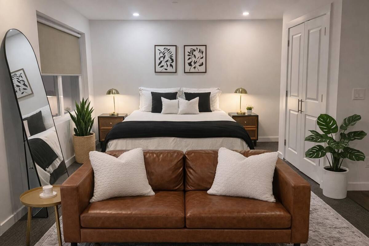Ein Schlafzimmer mit einem Bett, zwei Nachttischen mit Lampen, einem braunen Ledersofa, einem großen Spiegel und zwei an der Wand befestigten Kunstwerken.