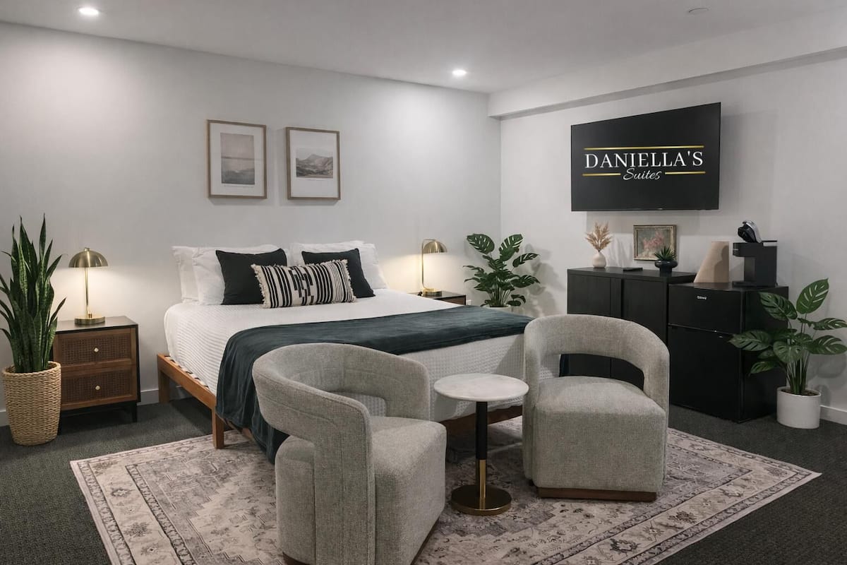 Ein modernes Schlafzimmer mit einem Bett, zwei Sesseln, einem kleinen Tisch, einem Nachttisch, einer Kommode und einem Fernseher, der "DANIELLA'S Suites" zeigt.