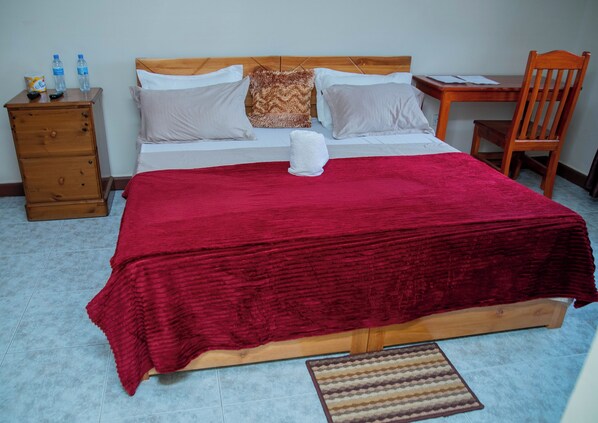 A neatly made bed with a red blanket, a wooden nightstand with remote controls, a wooden chair, and a small table with papers.