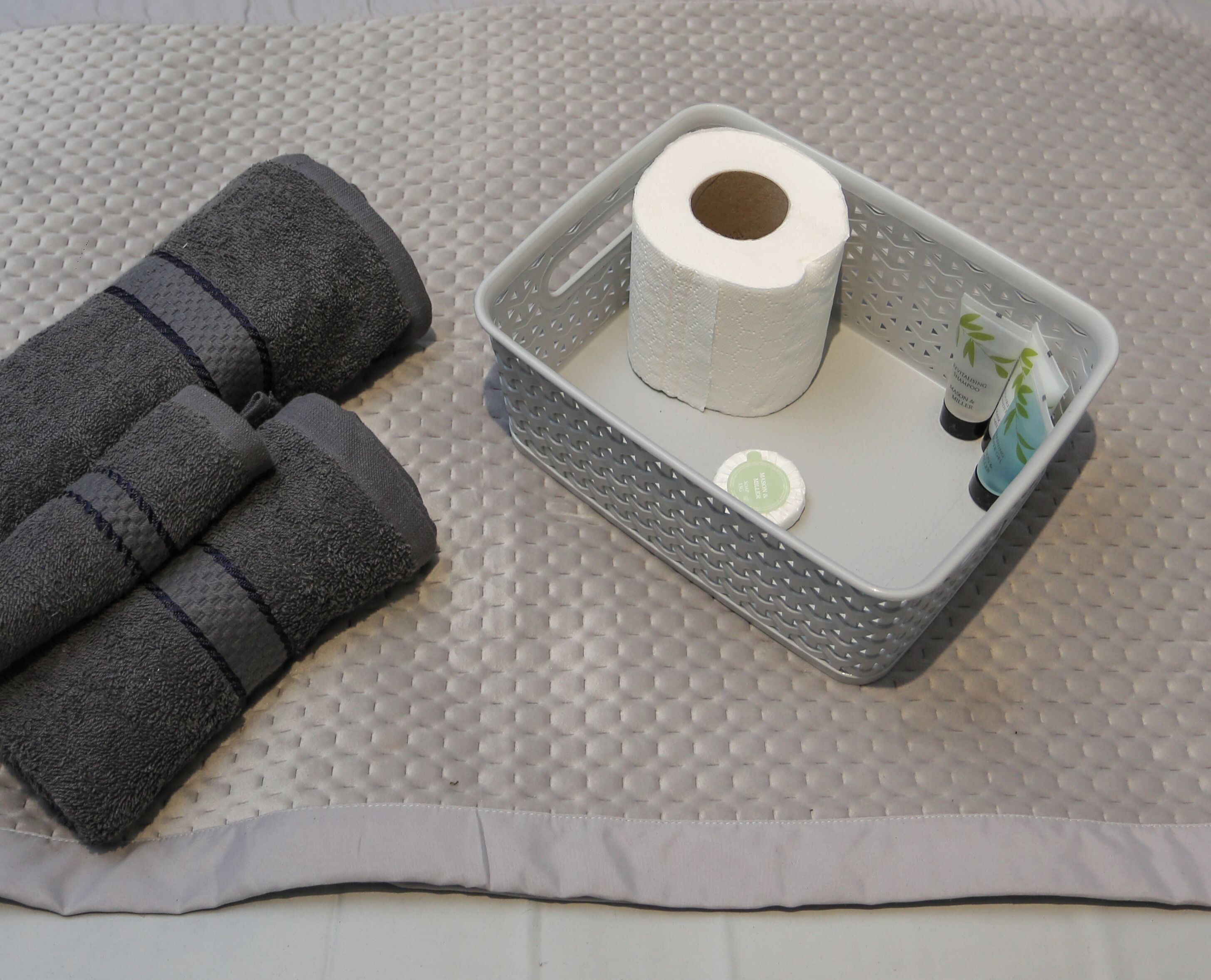 A bathroom with a textured floor, a gray towel, a roll of toilet paper, and a small bottle of hand soap.
