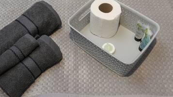A bathroom with a textured floor, a gray towel, a roll of toilet paper, and a small bottle of hand soap.