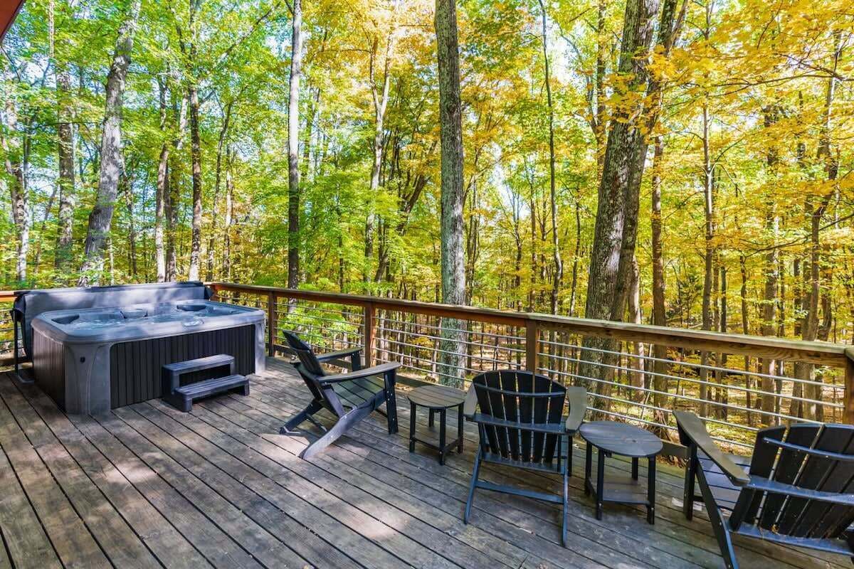 A deck with a hot tub, chairs, and tables overlooking a forest.