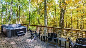 A deck with a hot tub, chairs, and tables overlooking a forest.