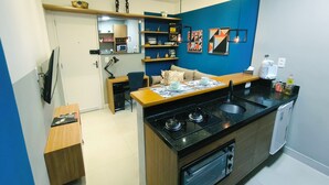 Een compacte keuken met een gasfornuis, een magnetron en een kleine koelkast. Er is een eethoek met een tafel en stoelen, plus een bank. De keuken heeft een blauwe accentmuur en planken met decoratieve objecten.