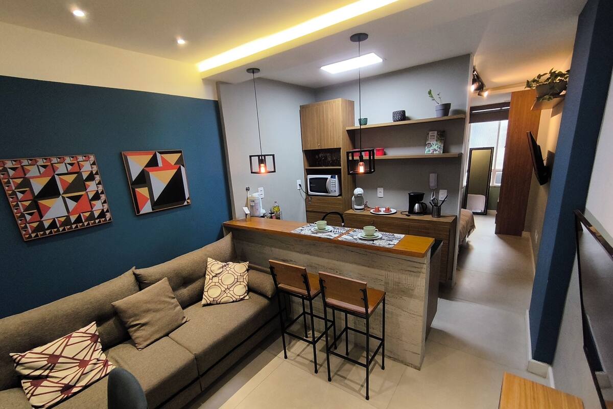 A modern living room with a sofa, a coffee table, and a bar area with stools. There is a kitchen in the background with a microwave, coffee machine, and shelves.