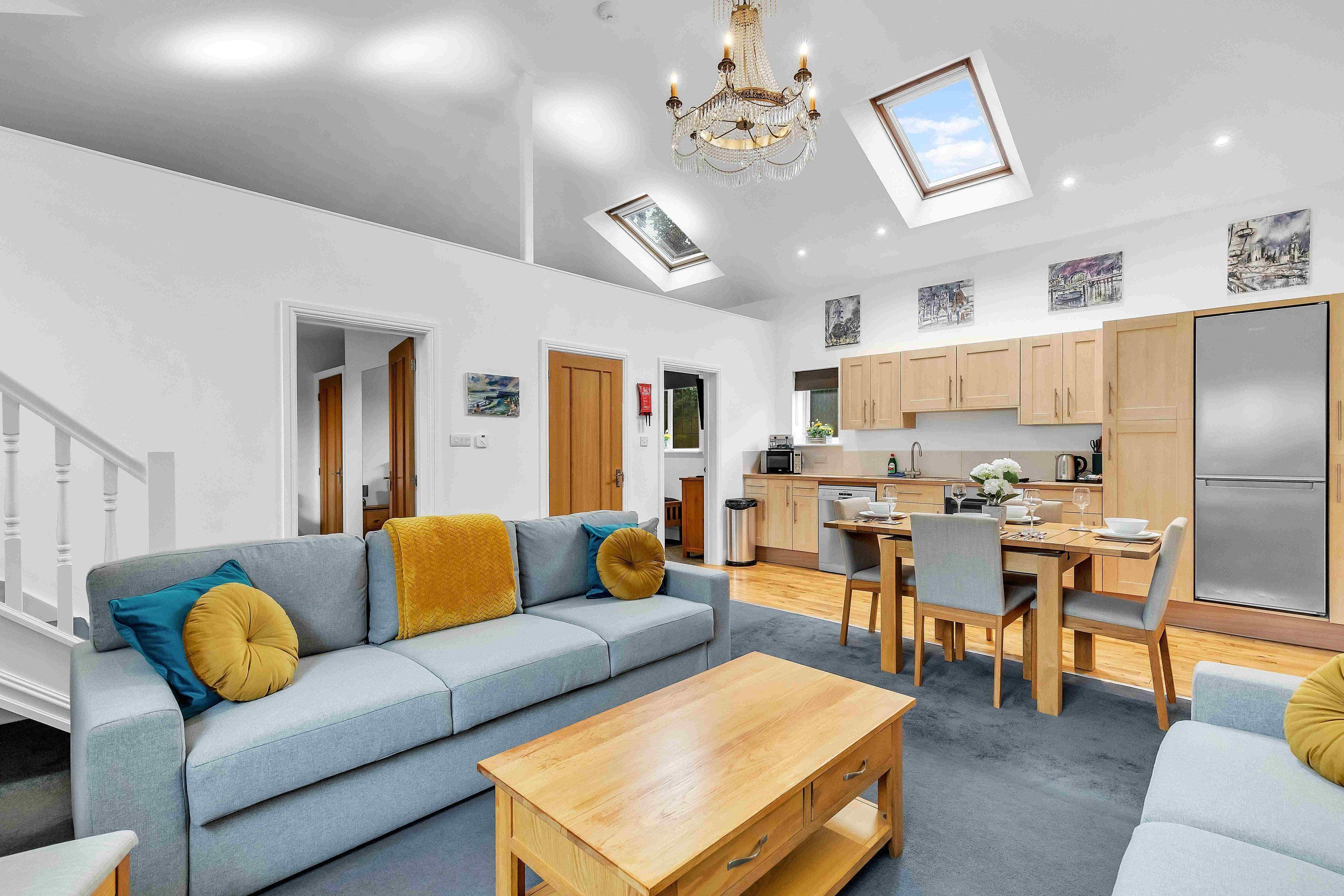 A modern living room with a sofa, coffee table, and dining area. There is a kitchen with wooden cabinets and a refrigerator.