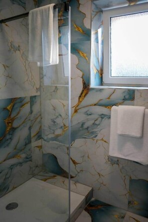 A bathroom with a glass shower enclosure, marble walls, and a window.