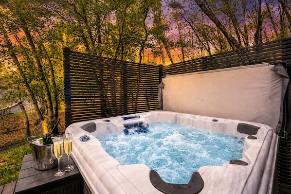 A hot tub with a wooden deck and a metal grill, surrounded by trees and a wooden fence.