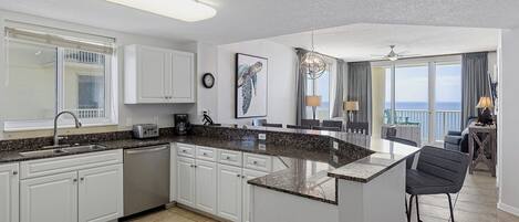 A modern kitchen with white cabinets, granite countertops, and a large island. There is a dining area with a view of the ocean.
