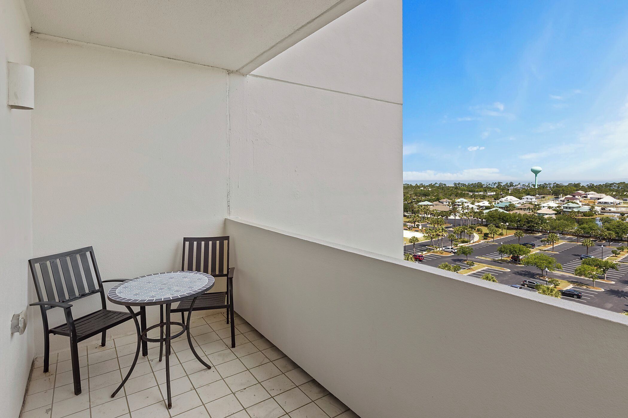 A balcony with a table and two chairs, overlooking a parking lot and buildings.