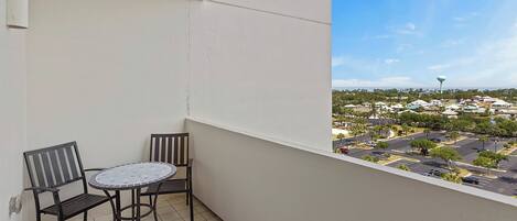 A balcony with a table and two chairs, overlooking a parking lot and buildings.