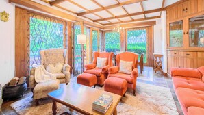 A spacious living room with wooden beams, a fireplace, and large windows offering a view of greenery.