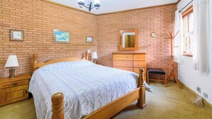 A bedroom with a brick wall, a wooden bed, a dresser, a mirror, a coat rack, and a window with curtains.