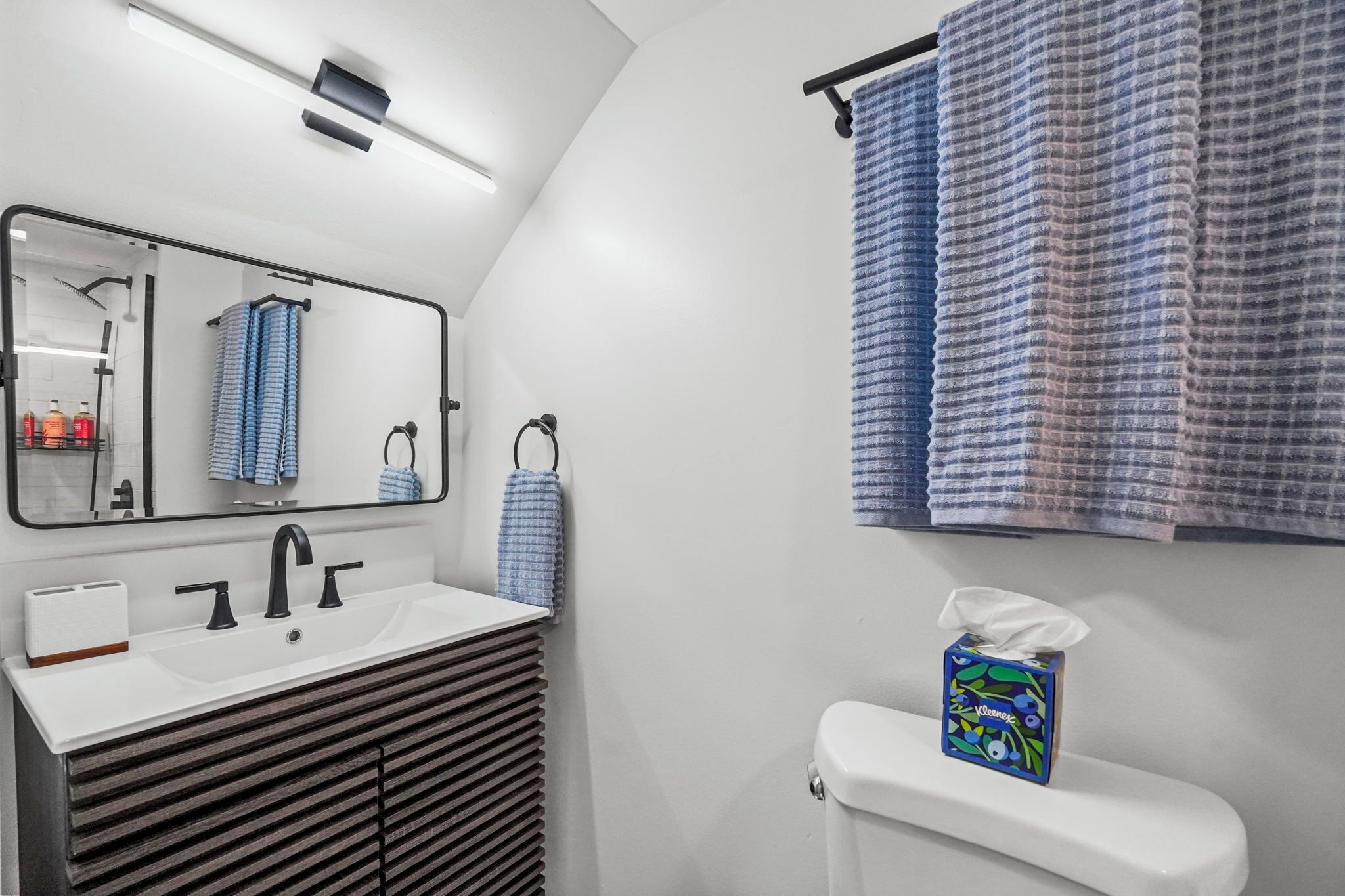 A modern bathroom with a white sink, black faucet, and a mirror above it. There are blue towels hanging on a rack, and a tissue box on the toilet.