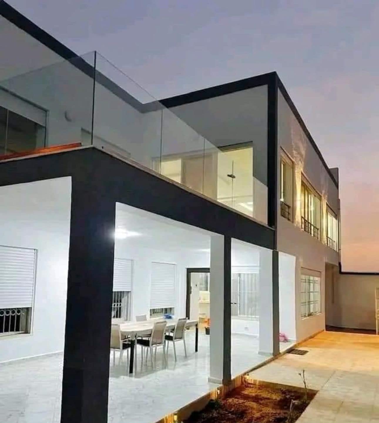 A modern two-story building with a glass facade, a flat roof, and a patio area with a dining table and chairs.