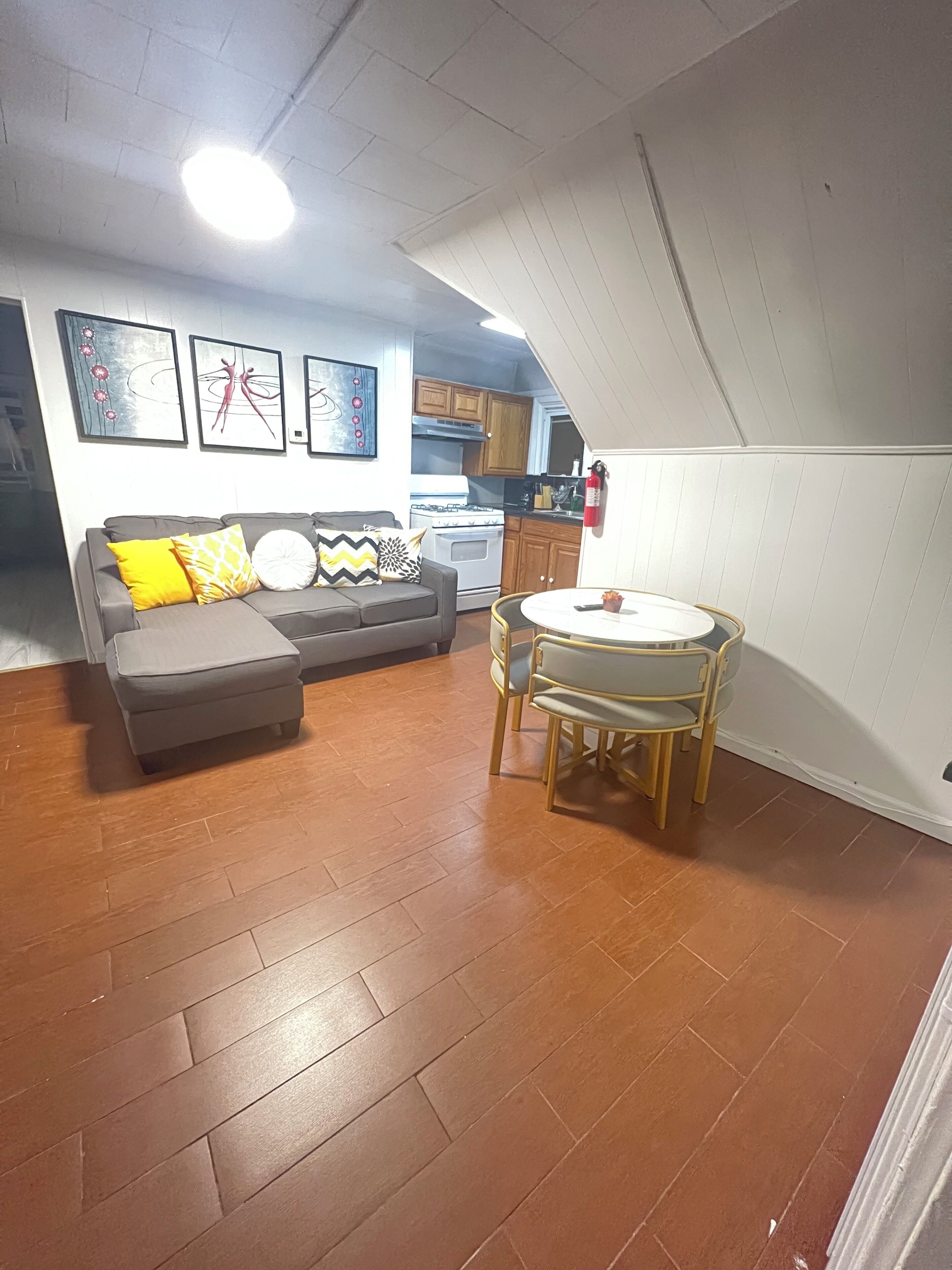 A compact living space with a sofa, ottoman, and a small dining table with chairs. A kitchen area with cabinets and a stove is visible in the background.