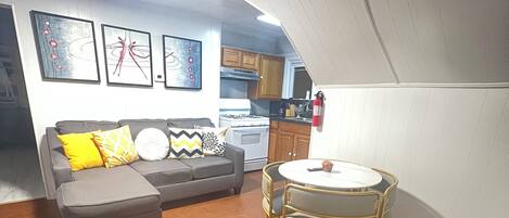 A compact living space with a sofa, ottoman, and a small dining table with chairs. A kitchen area with cabinets and a stove is visible in the background.