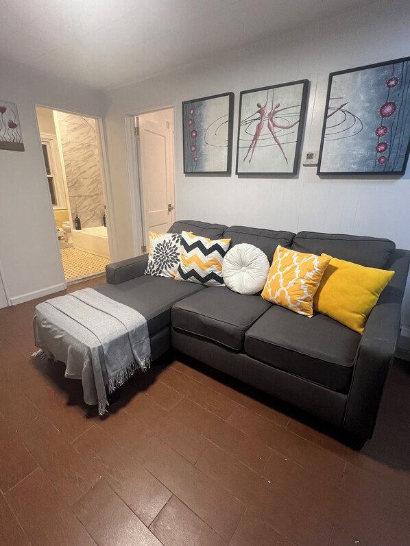 Ein Wohnzimmer mit einem grauen Ecksofa, einer grauen Decke und einem kleinen Hocker. An der Wand hängen drei gerahmte Kunstwerke.
