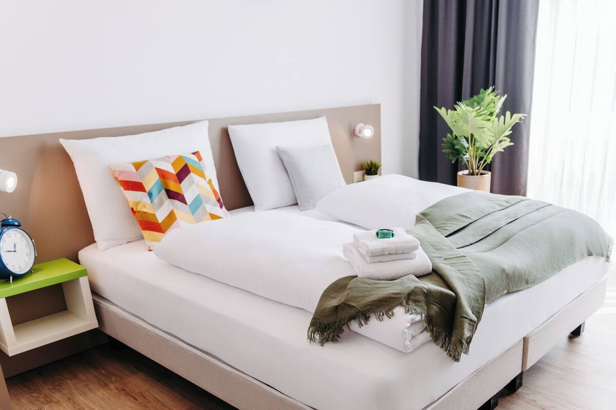 A neatly made bed with white linens, a colorful geometric pillow, and a green throw blanket.