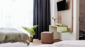Una habitación de hotel moderna con una cama, un escritorio pequeño con un espejo, un televisor montado en la pared y un ventanal con cortinas translúcidas.