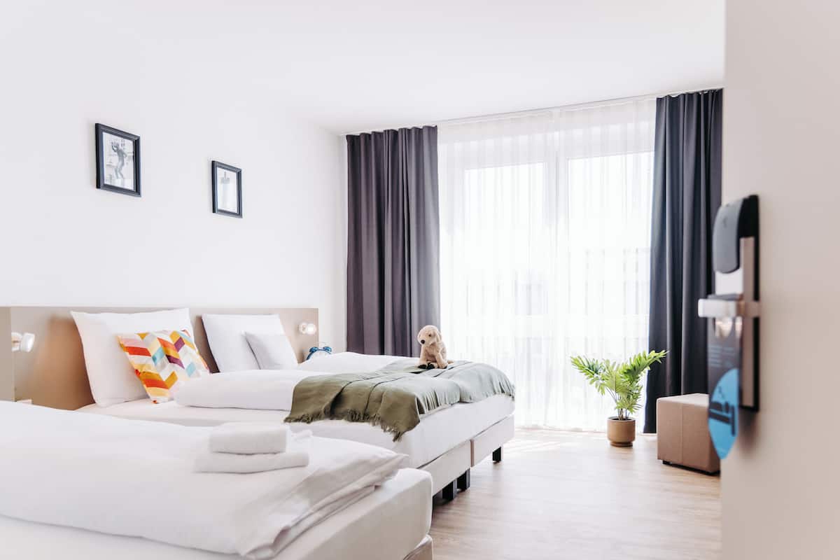 A modern hotel room with a large bed, a smaller bed, a sofa, a TV, and a potted plant.