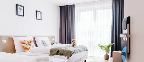 A modern hotel room with a large bed, a smaller bed, a sofa, a TV, and a potted plant.